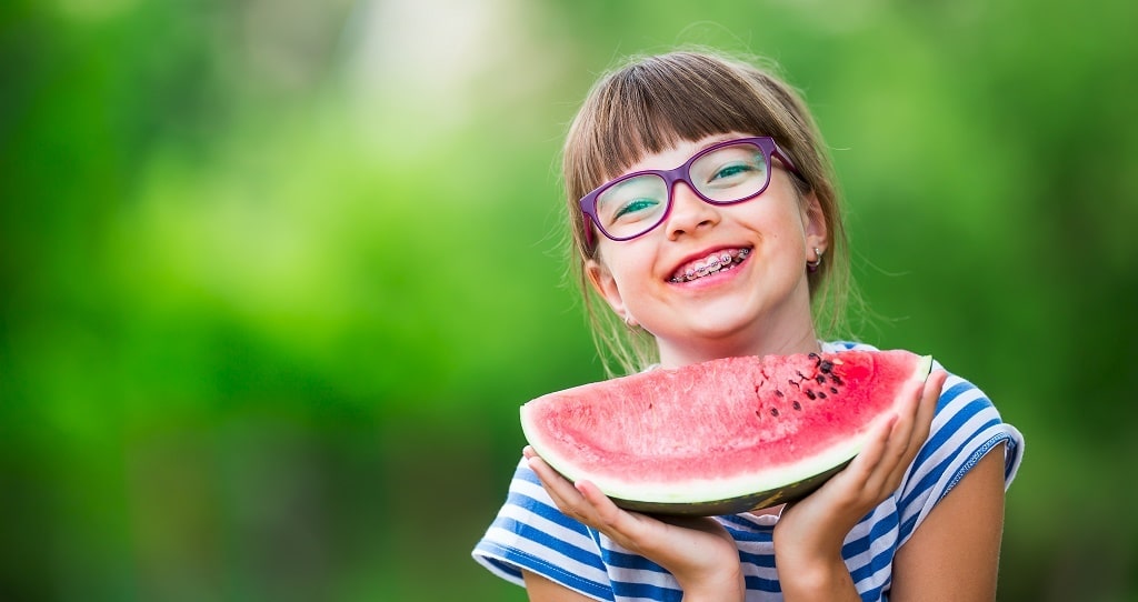 A little girl with meatl braces holding a watermelon which she can eat it with her braces on 