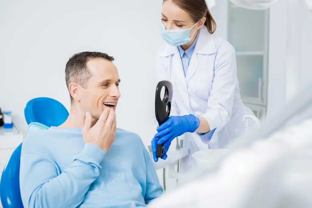 A dentist holding a mirror for a male patient looking at his teeth.